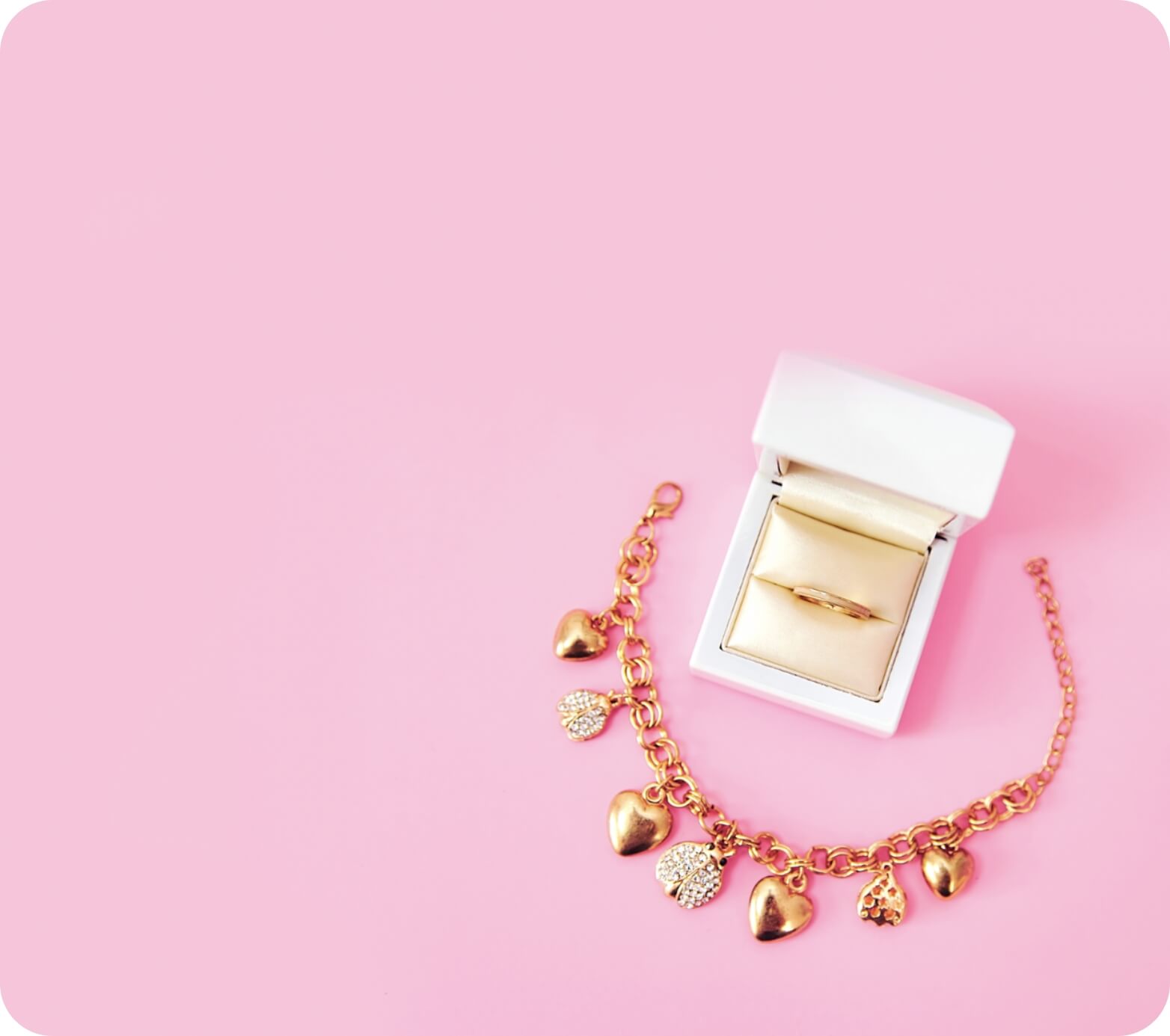 A golden heart charm bracelet and a ring in a white jewelry box, both placed on a pink background